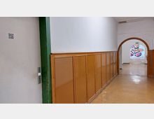 Ein schlichter Flur mit hellbraunem Holzvertäfelung an einer weißen Wand. Am Ende des Flurs befindet sich ein rundbogenförmiger Durchgang, hinter dem ein Wandgemälde mit bunten Motiven sichtbar ist.