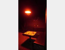 Ein dunkler Raum mit schwarzen Wänden und einer Sitzbank aus gepolstertem Kunstleder, die um einen kleinen quadratischen Tisch mit einer bunten, beklebten Oberfläche angeordnet ist. Eine einzelne, orangefarbene Hängelampe beleuchtet die Szene schwach.