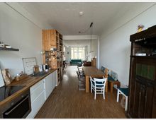 Wohnküche mit hellem Holzboden und weißen Wänden. Links befindet sich eine Küchenzeile mit Holzakzenten, daneben ein offenes Regal; rechts steht ein langer Esstisch mit verschiedenen Stühlen und Bänken. Im Hintergrund sind ein Wohnbereich mit Sofa und große Fenster zu sehen.