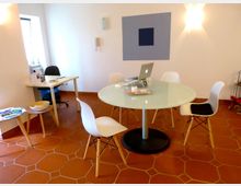 Ein heller Raum mit terrakottafarbenem Fliesenboden, ausgestattet mit einem runden Glastisch, umgeben von vier weißen Stühlen, sowie einem kleinen Arbeitstisch mit Bürostuhl in einer Ecke. An der Wand hängt ein abstraktes Kunstwerk, und durch ein Fenster fällt Tageslicht in den Raum.