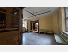 Ein leerer Raum mit gelb gestrichenen Wänden, einem gefliesten Boden und zwei großen Fenstern mit Gitterrahmen. Es gibt grüne Holzbänke entlang der Wände, eine offene Tür aus Glas und Metall führt zu einem angrenzenden Bereich, und eine rostige Metalltür ist teilweise sichtbar im Vordergrund.