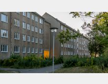 Mehrstöckiges Wohngebäude mit einer schlichten Fassade aus Putz, zahlreichen weißen Fenstern und einem offenen Eingangsbereich, der von innen beleuchtet ist. Der Vorplatz besteht aus einem Gehweg, wild wachsender Vegetation und Bäumen im Vordergrund.