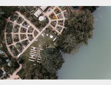 Luftaufnahme eines Gartens mit symmetrischen Blumenbeeten, geschwungenen Wegen und einer Rasenfläche. Im unteren Bereich sind gestellte Stühle und Tische zu sehen, rechts grenzt der Garten an ein Gewässer, umgeben von Bäumen.