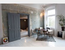 Esszimmer mit einem Holztisch und gemischten Stühlen auf einem gemusterten Teppich, graue Tür in einer abgeblätterten Wand, daneben ein großes Fenster mit weißen Vorhängen; modern-rustikale Gestaltung mit minimalistischen Details und Pflanzen.