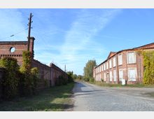 Straßenansicht eines verlassenen Industrie- oder Fabrikgeländes mit Backsteingebäuden auf beiden Seiten, teils von Pflanzen überwuchert. Der gepflasterte Weg verläuft zwischen den Gebäuden, und im Hintergrund sind Bäume und ein blauer Himmel sichtbar.