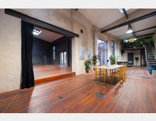 Geräumiger Industrieraum mit Holzboden, einer kleinen Bühne mit schwarzem Vorhang auf der linken Seite und einem Essbereich mit einem weißen Tisch und durchsichtigen Stühlen rechts. Im Hintergrund sichtbar: hohe Fenster, eine Metalltreppe zu einer Galerieebene, sowie Pflanzen und dekorative Elemente.