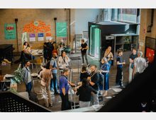 Menschen versammelt in einem offenen Raum mit Backsteinwänden, Stehtischen und Informationsplakaten an den Wänden. Im Hintergrund befinden sich große Fenster, eine grüne Notausgangstür und eine kleine Küchenecke.