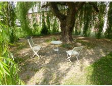 Ein kleiner Sitzbereich mit drei einfachen Gartenstühlen und einem runden Tisch aus Metall steht auf einem gepflasterten Boden unter einer großen Weide mit herabhängenden Zweigen. Im Hintergrund sind weitere Vegetation und eine Mauer sichtbar.