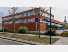 Backsteingebäude der Feuerwehr Luckenwalde mit großen Fenstern und der Beschriftung „Feuerwehr Luckenwalde 112“, umgeben von jungen Bäumen, Sträuchern und einem gepflasterten Gehweg; im Vordergrund ein Verkehrsschild und ein Straßenschild mit der Aufschrift „Hermann-Henschel-Weg“.