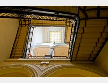 Blick von unten in ein mehrstöckiges Treppenhaus mit kunstvollen schwarzen Geländern und einem Oberlicht aus gelbem Glas. Die Wände sind cremefarben, mit dekorativen Bögen und verzierten Stuckelementen.