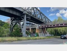 Ansicht einer stählernen Eisenbahnbrücke und einer Fußgängerbrücke in Berlin, umgeben von viel Grün. Unter der Brücke steht das Wort "Berlin" in großen Buchstaben auf gelben Backsteinsockeln, umgeben von Graffiti.