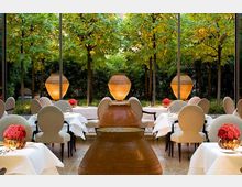 Elegantes Restaurant mit gedeckten Tischen und beigefarbenen Stühlen in einem hellen Raum mit großen Fenstern, durch die ein Garten mit Bäumen, Pflanzen und beleuchteten Keramikkrügen sichtbar ist. Die Tische sind mit roten Blumenvase-Dekorationen akzentuiert.