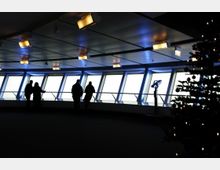 Innenraum einer Aussichtsplattform mit großen, schrägen Fenstern und bläulicher Beleuchtung; Personen stehen an einer Brüstung und ein geschmückter Weihnachtsbaum ist rechts im Vordergrund sichtbar.