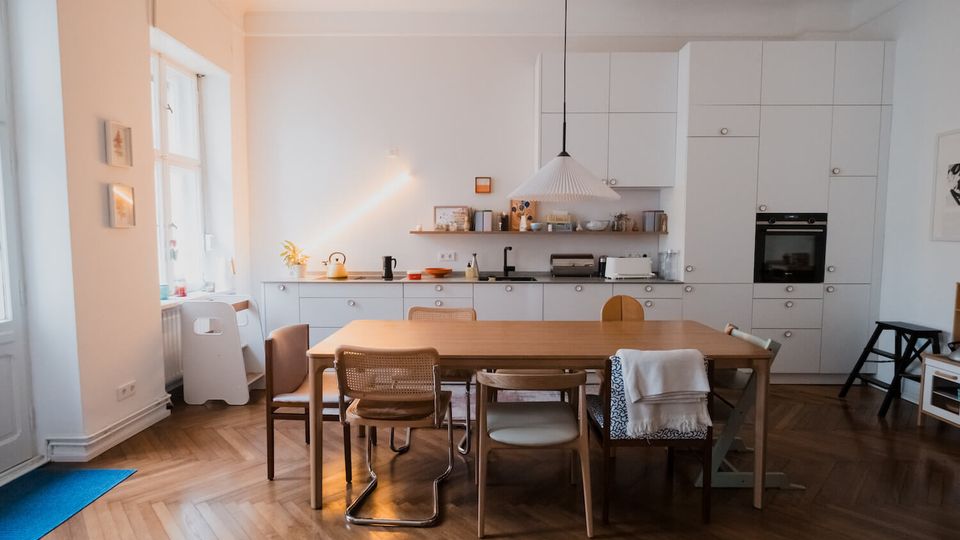 Bright open-plan kitchen and dining area with white built-in cabinets, a long countertop with sink and oven, and a large wooden table surrounded by mixed chairs. Tall windows on the left bring in natural light, and the room has a herringbone wood floor and a hanging pendant lamp over the table.