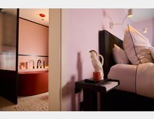 Modernes Schlafzimmer mit rosafarben gestrichenen Wänden, einem schwarzen Nachttisch mit einer weißen Keramikfigur und einer roten Kerze darauf. Im Hintergrund ist ein Badezimmer mit freistehender roter Badewanne, schwarzer Armatur und rosafarbenen Wänden sichtbar.