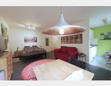 Geräumiges Studio-Apartment mit kombinierten Wohn- und Schlafbereichen. Im Vordergrund ein Holztisch unter einer großen, metallischen Hängelampe; im Hintergrund ein Bett, eine rote Couch, mehrere Bilder an den Wänden und eine offene Küche mit hellgrüner Wand.