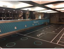 Ein moderner Fitnessraum mit einer schwarzen Trainingsfläche, die durch Linien und Kreise markiert ist, sowie der Aufschrift "Xpress". Im Hintergrund befindet sich eine blaue Wand mit geometrischem Muster und der Bezeichnung "Xpress", daneben diverse Fitnessgeräte und Beleuchtung an der Decke.