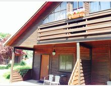 Zweistöckiges Holzhaus mit schrägem Dach und überdachtem Eingangsbereich. Der Balkon im Obergeschoss verfügt über Blumenkästen, und im Vordergrund stehen zwei Stühle neben einer kleinen Tischgruppe.