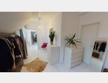 Ein heller, moderner Ankleideraum mit weiß gestrichenen Wänden und hellem Holzboden. Links befindet sich eine offene Kleiderstange mit verschiedenen Kleidungsstücken, rechts zwei weiße Kommoden mit Zimmerpflanzen darauf, ein langer Spiegel an der Wand, und ein kleiner Teppich in Felloptik auf dem Boden. Im Hintergrund führt eine Tür zu einem weiteren Raum mit Pflanzen.