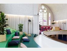 Ein moderner Innenraum in einer umgebauten Kirche mit hohen, spitzbogigen Fenstern und einer gewölbten Decke. Der Raum enthält stufenförmige Sitzbereiche in Grün und Rot, dekoriert mit Kissen und Pflanzen, wobei weiße Vorhänge entlang der Wände hängen.