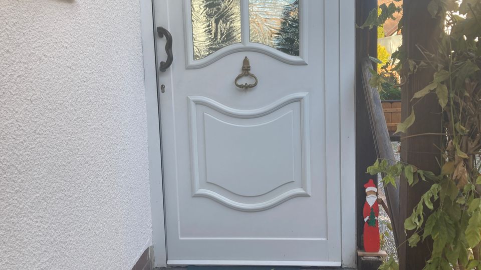 Weiße Haustür mit dekorativem Glasfenstereinsatz und einem roten Anhänger darüber, erreichbar über eine kleine Treppe mit dunklen Fliesen. Links ist eine verputzte Wand und rechts eine Holzstütze mit Pflanzen und weihnachtlicher Dekoration.