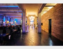 Großzügiger, modern eingerichteter Veranstaltungsraum mit hohen Decken, Betonpfeilern und großen Fensterfronten. Der Raum ist von warmem Licht beleuchtet, mit Holzwänden, schwarzen Tischen und Stühlen auf der linken Seite sowie Stehtischen in der Mitte.