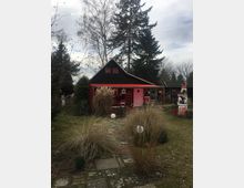 Kleines Holzhäuschen mit schwarzem Dach und rot gestrichener Veranda, umgeben von einem Garten mit Grasbüscheln, Gehwegplatten und Bäumen im Hintergrund. Der Himmel ist bewölkt, und es befinden sich dekorative Elemente wie Laternen und bunte Verzierungen vor dem Haus.