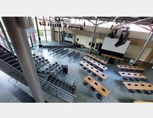 Ein moderner Veranstaltungsraum mit Bühne, Stuhlreihen und Tischen mit Mikrofonen. Im Vordergrund befindet sich eine Treppe, und an der Rückwand steht eine große Projektionsleinwand.