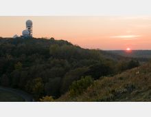 Eine Waldlandschaft auf einem Hügel mit der ehemaligen Abhörstation auf dem Teufelsberg in Berlin, erkennbar an Gebäuden mit großen, kuppelförmigen Radarkonstruktionen. Im Hintergrund geht die Sonne über dem leicht bewaldeten Horizont unter, der Himmel zeigt rötliche und orangefarbene Töne.