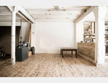 Heller, industrieller Raum mit Holzboden, sichtbaren weißen Säulen und einer weißen Backsteinwand; auf der linken Seite befinden sich Geräte und ein schwarzer Schrank, rechts ein Holztisch und Bücherregale im Hintergrund.