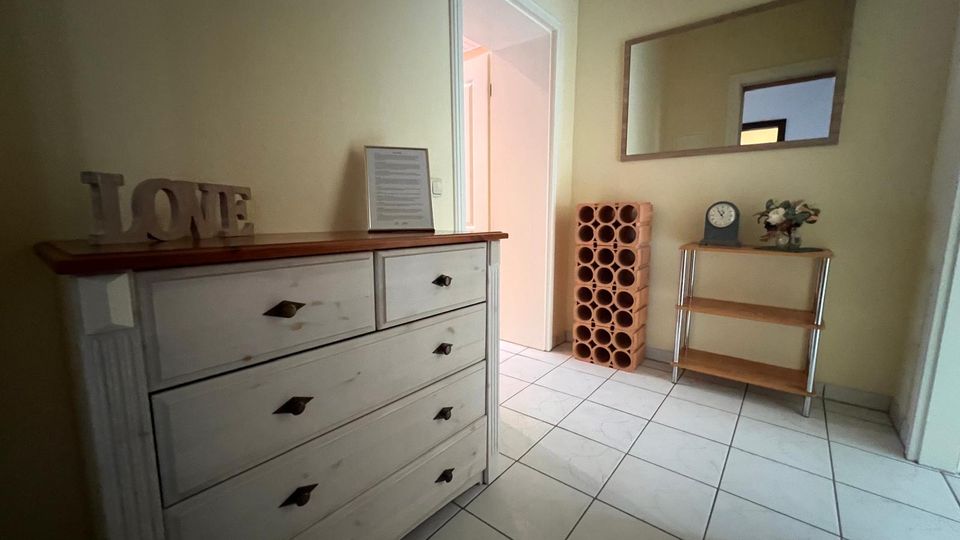 Heller Eingangs- oder Flurbereich mit weiß gefliestem Boden und cremefarbenen Wänden, ausgestattet mit einer weißen Kommode und einem offenen Durchgang zu einem Nebenraum. An der Wand hängen ein großer rechteckiger Spiegel; daneben stehen ein terrakottafarbenes Regal mit runden Fächern sowie ein kleines Holzregal mit Dekoration.