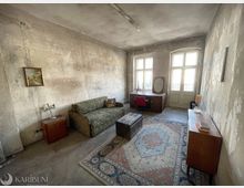 Großes, spärlich möbliertes Zimmer mit abgenutzten, unverputzten Wänden, einem gemusterten Sofa, Teppich und Holzmöbeln. Am Ende des Raums stehen zwei hohe Fenster mit einer verglasten Tür, die Tageslicht hereinlassen.