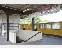 Bahnhof der Berliner U-Bahn mit gelbem U-Bahnzug der Linie U2 im Hintergrund. Der Bahnsteig verfügt über weiße Kachelwände, ein Treppenabgang und ein Stationsschild mit der Aufschrift "Ruhleben".