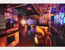 Industriehalle mit modernem, künstlerischem Ambiente, beleuchtet in bunten Farben. Im Raum befinden sich industrielle Strukturen, Fensterfronten, Galerien mit Treppen sowie ein großer gewölbter Luftschlauch mit farbiger Beleuchtung.