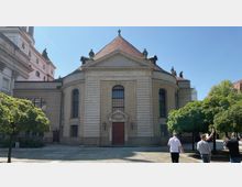 Sandsteinfassade einer barocken Kirche mit rotem Walmdach, hohen Rundbogenfenstern und einem reich verzierten Eingangsportal in der Mitte. Vor der Kirche steht ein kleiner Platz mit gepflastertem Boden und mehreren Bäumen, bei sonnigem Wetter.