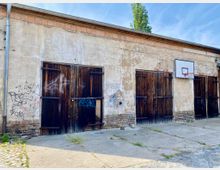 Eine alte, verputzte Lagerhalle mit Holz-Doppeltüren, teilweise mit Graffiti bedeckt. An der rechten Wand ist ein Basketballkorb montiert, und der Boden besteht aus Pflastersteinen und Betonflächen mit vereinzeltem Grasbewuchs.