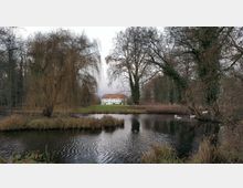 Eine Parklandschaft mit einem Teich im Vordergrund, umgeben von kahlen Bäumen und Gräsern. Im Hintergrund steht ein weißes Herrenhaus mit rotem Dach, eingebettet in eine weitläufige Wiese.