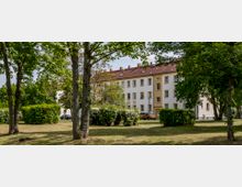 Mehrfamilienhaus im Stil eines Wohnblocks mit gelben und weißen Fassadenteilen sowie roten Ziegeldächern, umgeben von einer grünen Parklandschaft mit Bäumen, Sträuchern und gepflegten Grünflächen. Im Vordergrund sind mehrere Bäume und Büsche zu sehen.