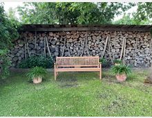Holzbank auf einer Rasenfläche vor einem überdachten Holzlager, in dem gestapelte Baumstämme und mehrere an der Wand hängende Äxte zu sehen sind. Daneben stehen zwei Pflanzkübel mit grünen Pflanzen, umgeben von Bäumen und Sträuchern.