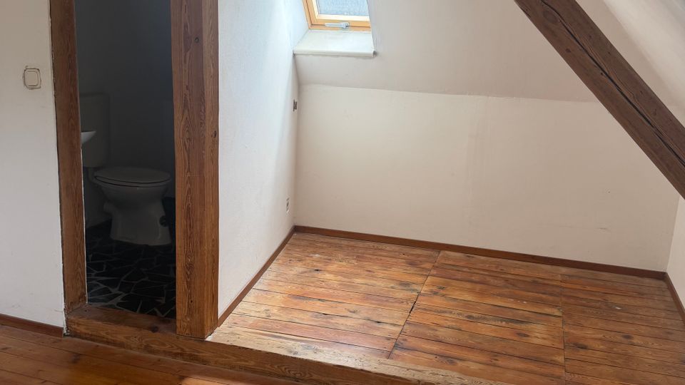 Dachgeschossraum mit schrägen Wänden, sichtbaren Holzbalken und Holzdielenboden; an der Wand sitzt ein kleines Dachfenster. Links führt ein offener Türrahmen in ein kleines Bad mit Toilette und dunklem Fliesenboden.