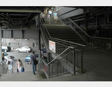 Treppenaufgang in einer städtischen Umgebung unter einer Brückenkonstruktion mit Metallträgern. Im Hintergrund sind parkende Autos und ein Backsteingebäude zu sehen, während Menschen die Treppe hinauf- und hinabgehen.