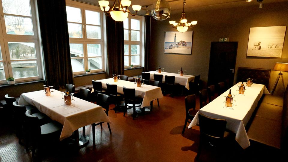 Innenraum eines Restaurants mit mehreren langen, weiß gedeckten Tischen und schwarzen Stühlen, beleuchtet von Deckenleuchten und einer Spiegelkugel. Große Fenster mit dunklen Vorhängen zeigen einen Blick auf Wasser und eine Brücke; an den grauen Wänden hängen gerahmte Bilder.