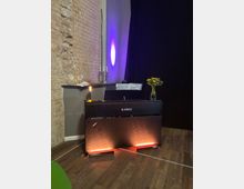 Alt text: A black upright digital piano sits on a wooden floor against an exposed brick wall and dark curtain backdrop, with purple uplighting on the wall. Sheet music is propped on the stand, and a small lamp and a vase of flowers rest on top of the piano.