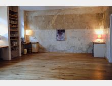 Raum mit rustikalem, verwittertem Putz an den Wänden, einem Holzboden und minimalistischer Einrichtung; links ein Regal mit Geschirr, rechts zwei kleine beleuchtete Tische und ein Kunstwerk an der Wand.