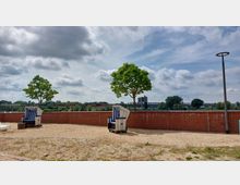 Sandfläche mit zwei Strandkörben vor einer niedrigen roten Ziegelmauer, dahinter eine grüne Landschaft mit Bäumen sowie einer Eisenbahnbrücke im Hintergrund. Der Himmel ist teilweise bewölkt, und eine Laterne steht am rechten Bildrand.
