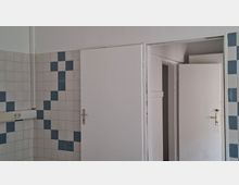 Innenraum mit weiß gekachelten Wänden und blauen Akzentfliesen im Schachbrettmuster sowie einer Steckdosenleiste an der Wand. Rechts sind zwei weiße Türen, eine davon geöffnet, die in einen angrenzenden Raum oder Flur führt.