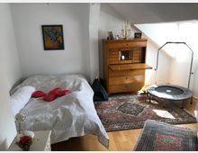 Ein kleines Schlafzimmer mit hellem Holzboden und schräger Zimmerdecke. Im Raum befinden sich ein Bett mit weißer Bettwäsche und roten Kissen, ein alter Holzsekretär, ein kleines Trampolin, ein Teppich mit orientalischem Muster und ein Beistelltisch mit Blumen.