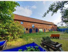 Großer ländlicher Backsteinbau mit einem roten Ziegeldach, blauen und grünen Fensterläden sowie Türen. Im Vordergrund sind eine Wiese, ein Gemüsebeet mit blauer Umrandung und ein orangefarbenes Sofa zu sehen, umgeben von dichtem Grün und Bäumen.