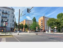 Straßenecke in einer urbanen Umgebung mit Mehrfamilienhäusern, darunter ein graues Gebäude mit einer großen Uhr und ein orangefarbenes Gebäude mit Balkonen. Im Vordergrund sind Ampeln, Fußgängerüberwege, Baustellenschilder und Bäume zu sehen.