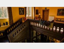 Ein Innenraum eines Hauses mit gelb gestrichenen Wänden, verziert mit Gemälden und Holzmöbeln. Im Vordergrund eine verzierte Holztreppe mit ovalen Ausschnitten, im Hintergrund Fenster mit Vorhängen, eine Tür und ein Beistelltisch.